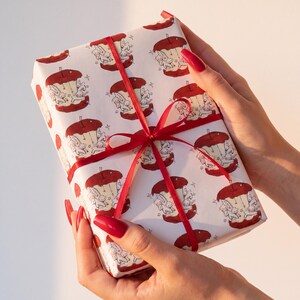 May include: A rectangular gift box wrapped in white paper featuring a repeating pattern of red apple cores with small white creatures inside. The package is tied with a thin, vibrant red satin ribbon arranged in a cross pattern with a central bow.