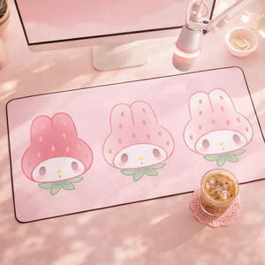 May include: A pink rectangular mousepad featuring three cartoon characters with strawberry-shaped hats. The characters have white faces, small eyes, and green leaves. The mousepad is on a pink desk with a computer monitor and a microphone.