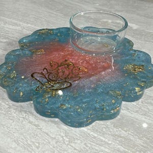 May include: A decorative resin coaster with a clear glass votive holder. The coaster is blue and red with gold flecks and a gold teapot design. The coaster has a scalloped edge.