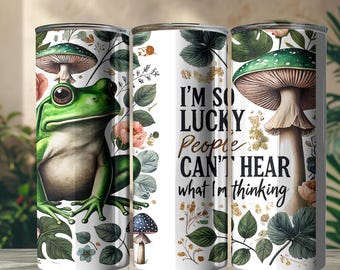 May include: A white tumbler featuring a green frog, mushrooms, and floral elements. The tumbler has the text "I'm so lucky people can't hear what I'm thinking" in black and gold lettering. The design includes leaves and flowers.
