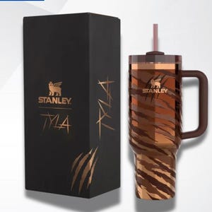 Puede incluir: Un vaso Stanley Tyla con un dise&ntilde;o de rayas de tigre marr&oacute;n y beige, un asa marr&oacute;n y una pajita a juego. Junto a &eacute;l, una caja negra con el logotipo de Stanley y el nombre "Tyla" en letras doradas.
