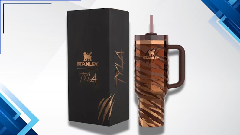 Puede incluir: Un vaso Stanley Tyla con un dise&ntilde;o de rayas de tigre marr&oacute;n y beige, un asa marr&oacute;n y una pajita a juego. Junto a &eacute;l, una caja negra con el logotipo de Stanley y el nombre "Tyla" en letras doradas.