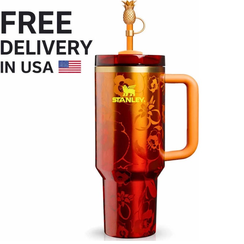May include: A tall, cylindrical Stanley tumbler with a metallic red and orange gradient finish featuring a floral pattern. It has a sturdy orange handle, a gold-toned rim, and a straw topped with a small gold pineapple-shaped charm.