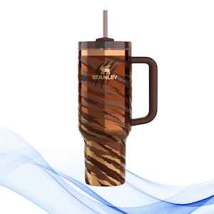 Puede incluir: Un vaso Stanley con un dise&ntilde;o de rayas de tigre marr&oacute;n. El vaso tiene un asa, una pajita y el logotipo de Stanley. El vaso est&aacute; dise&ntilde;ado para mantener las bebidas fr&iacute;as o calientes. Es una botella de agua reutilizable.