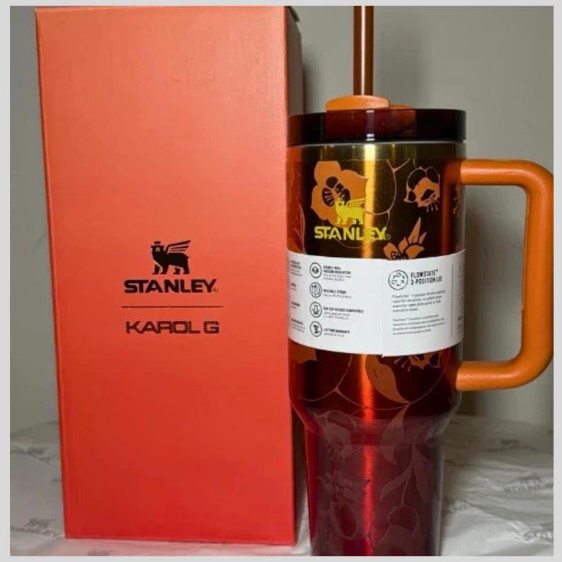 May include: A metallic orange and red Stanley tumbler with a floral pattern, a matching orange handle, and a straw. The tumbler features a white product label and is positioned next to a tall orange box labeled Stanley and Karol G with a winged bear logo.