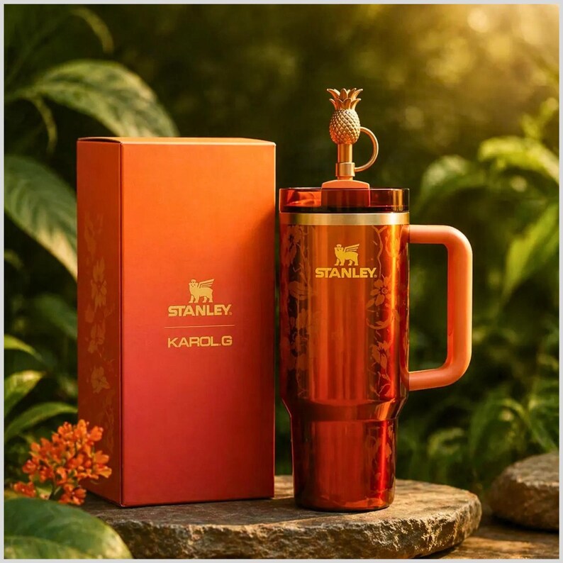 May include: A metallic orange Stanley tumbler with a floral pattern, a matching handle, and a gold-toned pineapple-shaped topper on the lid. Next to it stands a rectangular orange box featuring the Stanley logo and the text KAROL G in gold.