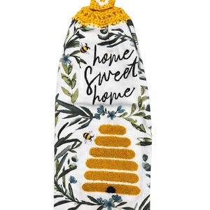 May include: White kitchen towel with a yellow crocheted top and a floral design. The towel features the words "home sweet home" and a yellow beehive illustration with bees. The towel is decorated with green leaves and blue flowers.