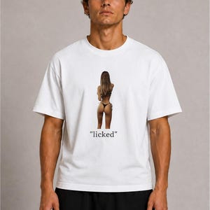 Könnte beinhalten: Ein weißes kurzärmeliges T-Shirt mit einem zentrierten Fotodruck einer Person von hinten, die einen schwarzen Bikini trägt. Unter dem Bild ist das Wort "licked" in einer schwarzen Serifenschrift gedruckt. Das T-Shirt hat einen Rundhalsausschnitt.