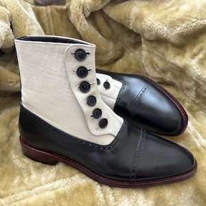 Puede incluir: Botines de dos tonos con puntera y talón de cuero negro y parte superior de tela crema. Las botas tienen cierre de botones y suela burdeos. Se exhiben sobre una superficie texturizada de color marrón dorado. Calzado elegante.