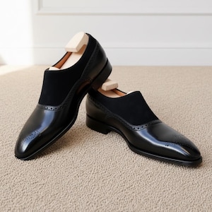 Side angle of mens black formal slip-on shoes showing buckle detail and brogue perforations