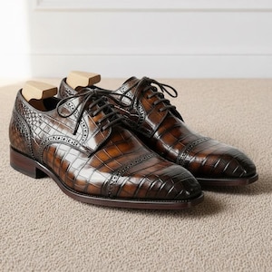 May include: A pair of brown leather dress shoes with a crocodile pattern. The shoes have a classic Oxford design with laces and wooden shoe trees inside. The leather has a glossy finish and a dark brown sole.