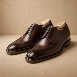 May include: A pair of brown leather dress shoes with a wingtip design. The shoes have black laces and are displayed with wooden shoe trees inside. The leather has a polished, glossy finish.