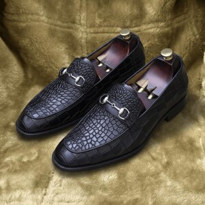May include: A pair of black leather loafers with a crocodile texture. Each shoe features a silver-colored metal ornament and a wooden shoe tree inside. The shoes are displayed on a gold-colored, textured surface.