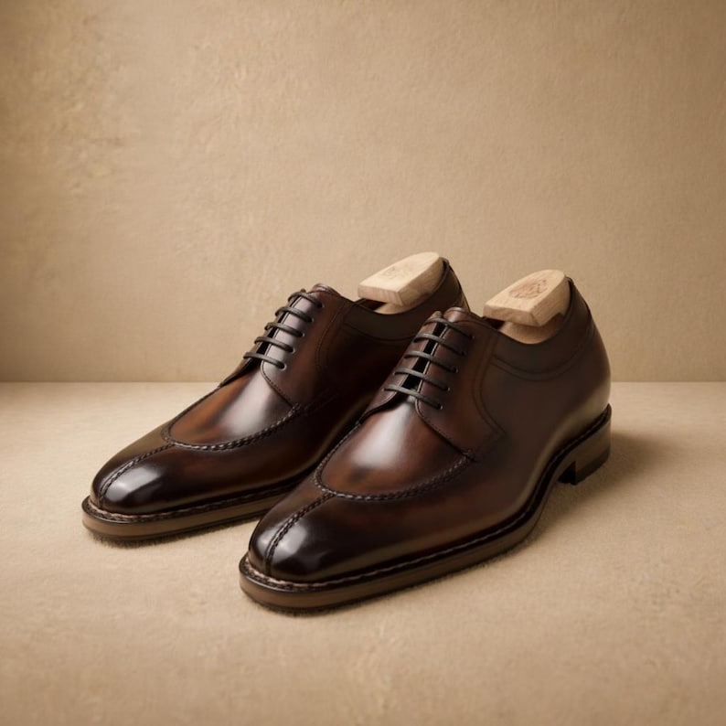 May include: A pair of brown leather dress shoes with a wingtip design. The shoes have black laces and are displayed with wooden shoe trees inside. The leather has a polished, glossy finish.