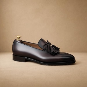 May include: A dark brown leather loafer with a tassel detail. The shoe has a rounded toe and a low heel. The shoe is displayed on a neutral background.