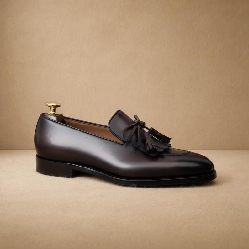 May include: A dark brown leather loafer with a tassel detail. The shoe has a rounded toe and a low heel. The shoe is displayed on a neutral background.