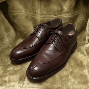 May include: A pair of brown leather dress shoes with a classic design. The shoes feature a lace-up closure, a rounded toe, and decorative stitching. The shoes are displayed on a soft, textured surface.