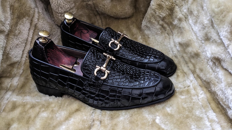 Black crocodile embossed leather loafers with metal horsebit on plush fabric background