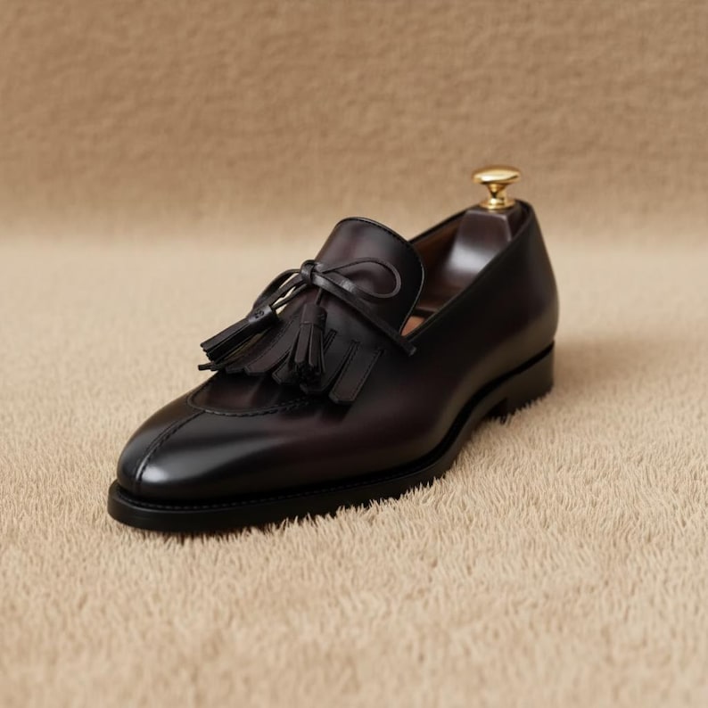 Men's dark brown smooth leather tassel loafer with kiltie fringe and leather bow detail, elevated three-quarter front view on a beige textured surface with wooden shoe tree visible
