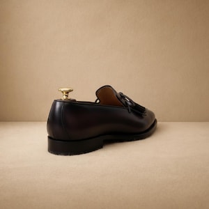 Close-up of handcrafted leather tassel and kiltie fringe on a dark brown men's slip-on loafer, showing leather bow tie accent and moc-toe stitching