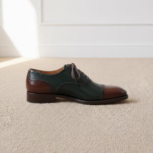May include: A two-tone leather oxford shoe. The shoe features a dark green upper, a brown heel and toe cap, and brown laces. The shoe is designed with decorative perforations along the seams.