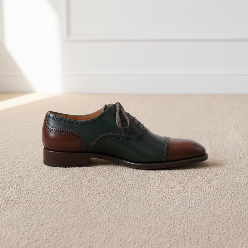 May include: A two-tone leather oxford shoe. The shoe features a dark green upper, a brown heel and toe cap, and brown laces. The shoe is designed with decorative perforations along the seams.