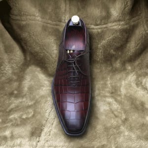 May include: A burgundy leather dress shoe with a textured, crocodile-like pattern. The shoe has a pointed toe, a lace-up closure, and a wooden shoe tree inside. The shoe is displayed on a soft, brown surface.