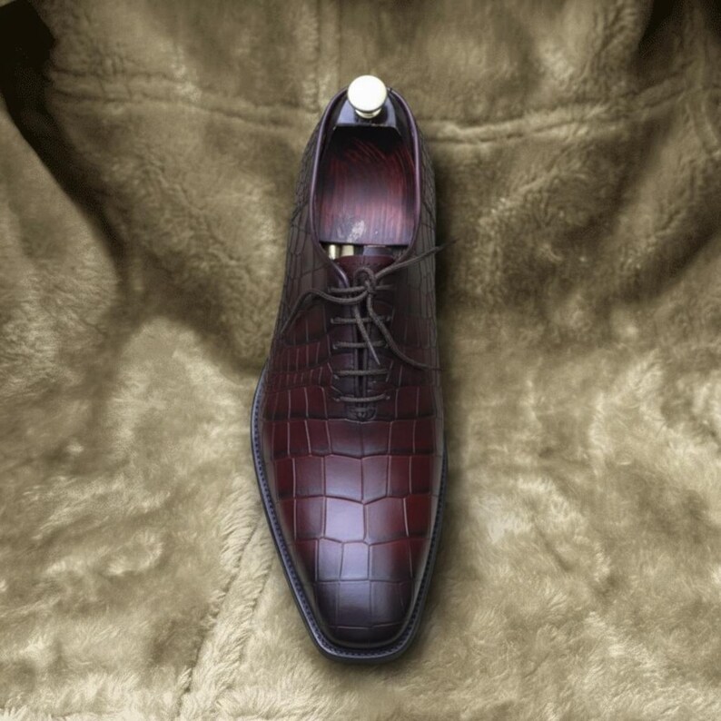 May include: A burgundy leather dress shoe with a textured, crocodile-like pattern. The shoe has a pointed toe, a lace-up closure, and a wooden shoe tree inside. The shoe is displayed on a soft, brown surface.