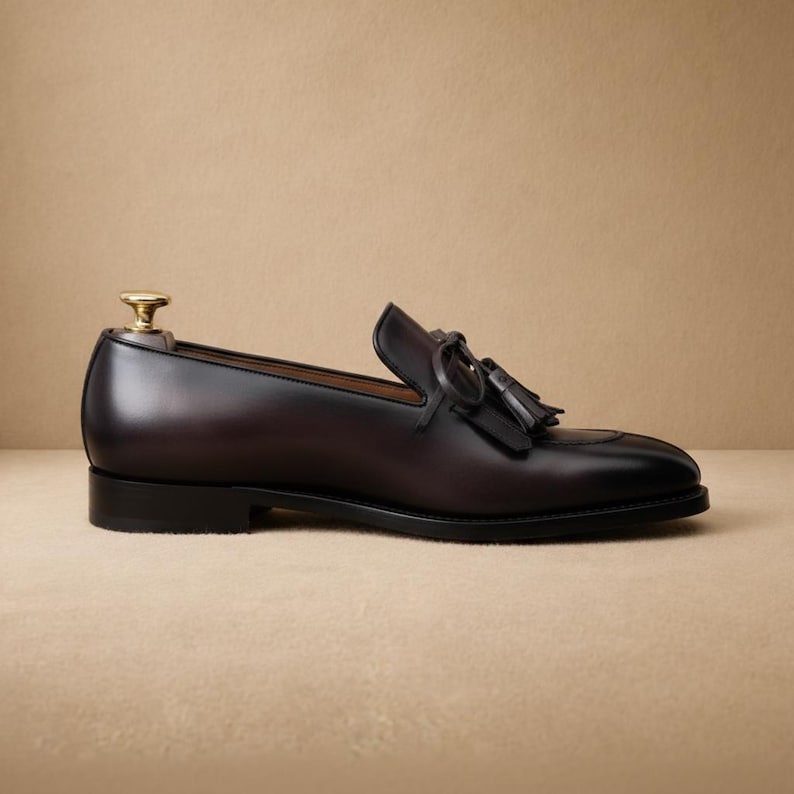 Side profile of men's dark brown tassel loafer showing smooth leather upper, black leather outsole, low heel stack, and pointed moc-toe silhouette