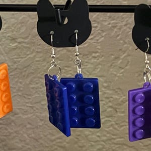 May include: A collection of colorful, square-shaped earrings in yellow, orange, blue, purple, and green. Each earring is designed to resemble a building block, with a silver hook for pierced ears. The earrings are displayed on a black rack.