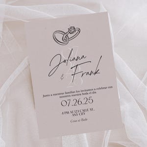 May include: A light pink wedding invitation with a ring illustration at the top. The names "Juliana & Frank" are written in elegant script. The date "07.26.25" and address are also included.
