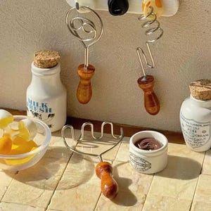 Könnte beinhalten: Miniatur-Küchenutensilien und Zubehör, darunter ein Schneebesen, eine Gabel und ein Schokoladentopf. Die Werkzeuge haben Holzgriffe und hängen an einer weißen Oberfläche. Eine kleine Glasschale enthält Eierschalen und Eigelb. Eine kleine weiße Flasche mit einem Korken ist ebenfalls vorhanden.