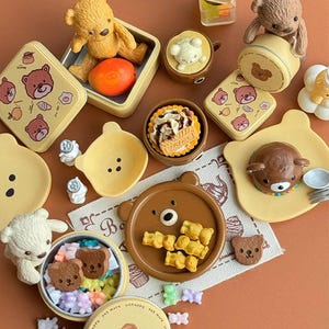 以下が含まれることがあります： クマをテーマにしたミニチュアアイテムのコレクション。お皿、容器、置物など。アイテムは茶色と黄色の色合いで、小さな食品が入っているものもあります。全体的な美しさは可愛らしくて気まぐれです。