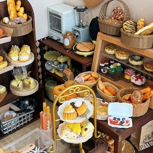 Könnte beinhalten: Miniatur-Bäckerei-Szene mit verschiedenen Backwaren, darunter Brot, Gebäck und Kuchen. Ausgestellt auf Regalen, in Körben und auf einem mehrstöckigen Ständer. Enthält einen Miniatur-Backofen, einen Mixer und einen Waffeleisen. Die Szene ist in einer warmen, einladenden Umgebung.