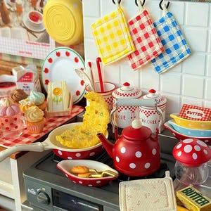 May include: A miniature kitchen playset featuring a red polka-dot kettle, a frying pan with a yellow omelet, two canisters labeled Sucre and Chicoree, a mushroom-shaped jar, a plate with strawberry motifs, and various small food items and linens.