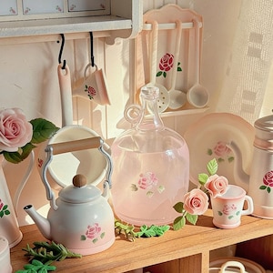 May include: A miniature kitchen scene in white and pink, featuring a kettle, water jug, mug, and utensils, all decorated with rose designs. The scene is set on a wooden shelf, evoking a vintage aesthetic.