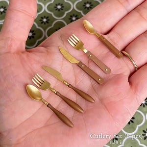 May include: A miniature cutlery set with six pieces, including spoons, forks, and knives. The utensils have gold-colored blades and brown handles. The text "Cutlery Set (6 pcs) 3.5 cm" is visible.
