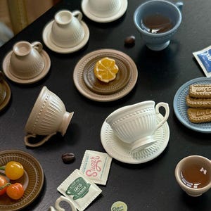 Puede incluir: Un surtido de tazas de té, platillos y platos de cerámica en miniatura en varios colores, incluyendo blanco, beige y azul. Algunas tazas contienen líquido. La escena incluye pequeños paquetes de azúcar y café, y una rodaja de limón.