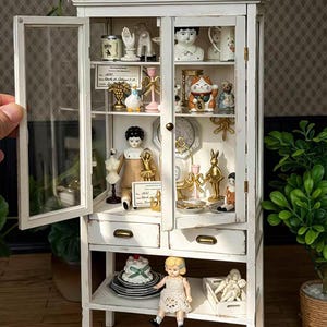 Peut inclure: Une vitrine miniature en bois blanc avec des portes vitrées et deux tiroirs, remplie de diverses petites figurines décoratives, dont une poupée, une statue de chat, un sablier et des meubles miniatures, disposés sur plusieurs étagères.