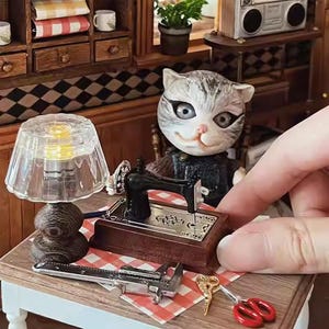May include: A detailed miniature sewing room scene featuring a cat figurine, a sewing machine, and a lamp. The scene includes a miniature sewing machine, a pair of scissors, and a caliper. The cat figurine is gray and white.