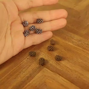 Puede incluir: Un conjunto de pequeñas cuentas decorativas con forma de piña. Algunas cuentas se sostienen en una mano, mientras que otras están esparcidas sobre una superficie de madera. Las cuentas tienen una base marrón con detalles blancos.