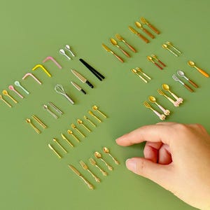 May include: Miniature cutlery set with various spoons, forks, knives, and chopsticks in gold and silver tones. Includes a whisk and colorful straws, all arranged on a green surface. A hand is reaching towards the objects.