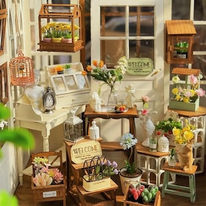 May include: Miniature garden scene with various decorative elements. Features small wooden structures, potted plants, angel figurines, and a "WELCOME" sign. The scene is set against a white door with a window.