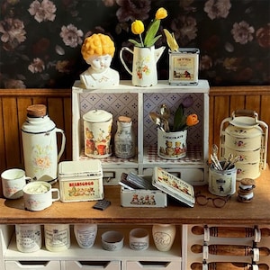 Puede incluir: Una colección de artículos de cocina vintage en miniatura, que incluye un busto, una tetera con flores amarillas y varias latas y tarros. Los artículos están dispuestos en una estantería de madera con un fondo de pantalla floral. Los artículos están pintados en crema y blanco con diseños florales y de osos de peluche.