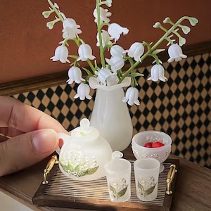Op de afbeelding: Miniatuur wit porseleinen theeservies met een dienblad, een vaas met witte bloemen en een kleine kom met rode objecten. De theeset bevat een theepot, suikerpot en twee kopjes, allemaal versierd met een bloemmotief. Het dienblad heeft goudkleurige handvatten.