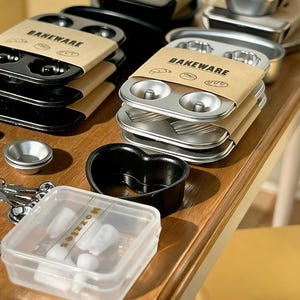 May include: A selection of metal bakeware, including muffin tins, doughnut pans, and a heart-shaped mould. The items are packaged with brown paper bands that read "BAKEWARE". A clear plastic container with the word "Nozzles" is also visible.