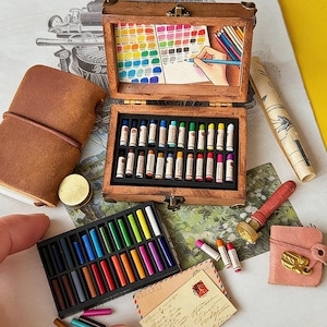 Può includere: Una scatola di legno in miniatura per forniture artistiche contenente due file di piccoli pastelli, con un'illustrazione sul coperchio interno di una mano che disegna. Include un taccuino in pelle marrone, un vassoio di pastelli, un timbro e una busta.