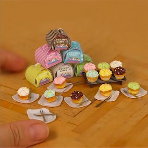 Puede incluir: Miniaturas de cupcakes con glaseados coloridos y cucharas diminutas se disponen sobre pequeños platos blancos. Varias cajas pequeñas estampadas se apilan detrás de los cupcakes. Las cajas tienen el texto "CREAMY DAILY".