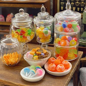 Può includere: Una collezione di barattoli in miniatura in vetro trasparente e piattini in ceramica bianca riempiti di caramelle colorate. I barattoli contengono orsetti gommosi, caramelle rotonde e lecca-lecca a strisce. I piattini contengono piccole caramelle arancioni a forma di frutto e lecca-lecca rotondi.