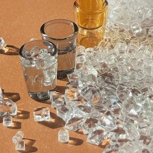 May include: Clear shot glasses filled with ice cubes, alongside a pile of clear, faceted ice-like crystals. The glasses are on a warm, light brown surface, with a few amber-colored shot glasses in the background.